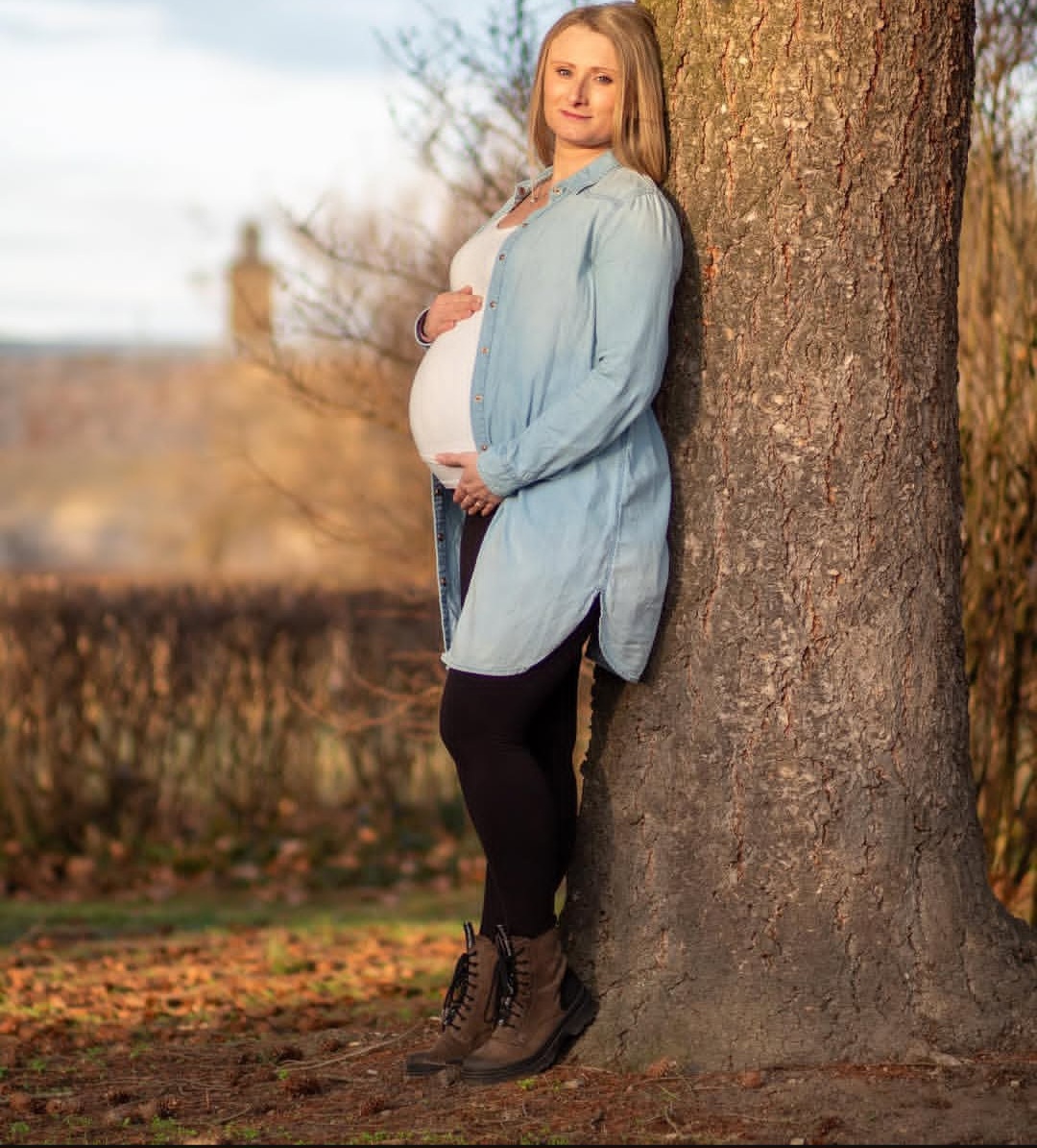 Těhotná žena v modré košili opřená o strom - pohled zepředu