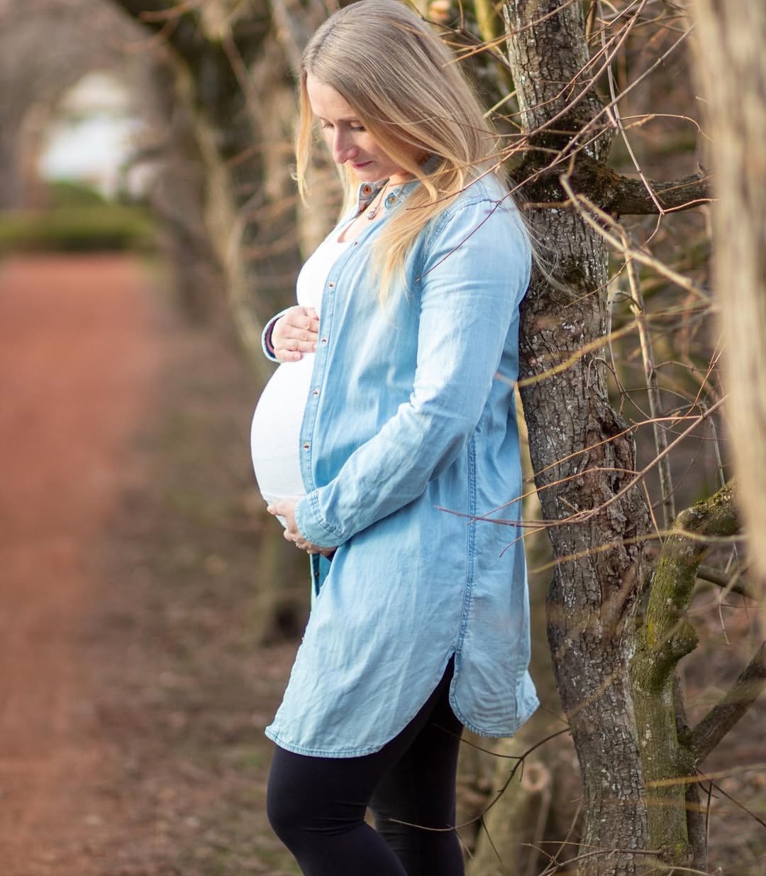 Těhotná žena v modré košili opřená o strom - boční pohled s bříškem